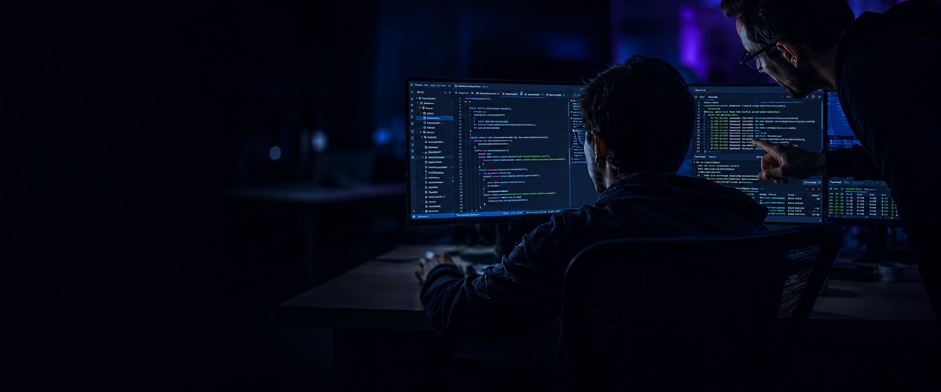 Two Telebridge developers in a dark office during a late code review, one pointing at a pull request across dual monitors